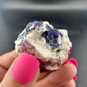 Pode incluir: Close-up de um espécime mineral natural com uma mistura de cores. A rocha tem uma base de tons esbranquiçados e castanho-avermelhados, com manchas marcantes de cristais azul escuro. O mineral é segurado em uma mão com esmalte rosa brilhante.