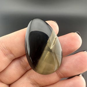 Könnte beinhalten: Ein polierter, tropfenförmiger Edelstein, diagonal in zwei Abschnitte geteilt. Eine Seite ist glänzend schwarz, die andere schimmernd goldbraun. Der Stein wird in einer Hand vor dunklem Hintergrund gehalten, wodurch seine reflektierenden Oberflächen hervorgehoben werden.