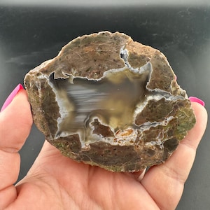 May include: A polished agate stone slice with a central cavity. The stone features bands of brown, white, and gray, with a dark brown outer edge and a hint of green. The stone is held in a hand.