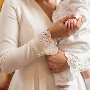 Può includere: Una persona in un vestito bianco tiene in braccio un bambino in un completo bianco. L'abito ha maniche lunghe con polsini in pizzo e bottoni di perle. Il bambino indossa un completo bianco con un colletto a balze.
