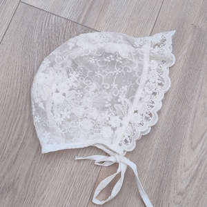 Op de afbeelding: Een witte kantmuts met een delicaat bloemenpatroon en een gebonden lint.