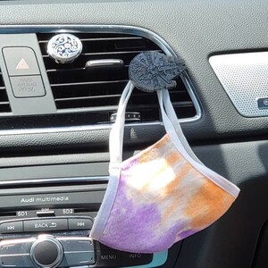 Puede incluir: Una mascarilla facial de tie-dye colgando de una ventilación de aire del coche con un ambientador de coche Millennium Falcon de Star Wars. Los controles de la radio del coche son visibles debajo de la ventilación.