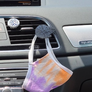 以下が含まれることがあります： 車のエアコンの吹き出し口に、紫とオレンジのタイダイ柄のマスクがぶら下がっています。吹き出し口には、ミレニアム・ファルコンの小さな黒い3Dプリントモデルが取り付けられています。