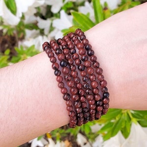May include: A stack of six bracelets made of small, round, reddish-brown beads with black inclusions. The bracelets are worn on a person's wrist, with a blurred background of green foliage and white flowers.