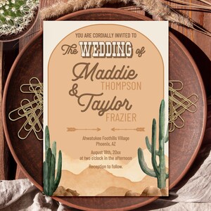 May include: A rectangular wedding invitation card featuring a tan background with an arched design, green watercolor cacti, and brown desert hills. The text reads: You are cordially invited to the wedding of Maddie Thompson and Taylor Frazier.
