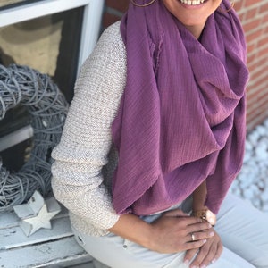 Può includere: Una sciarpa di cotone viola con un bordo sfrangiato. La sciarpa è drappeggiata intorno al collo e alle spalle di una persona.