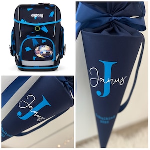 Könnte beinhalten: Marineblauer Schulrucksack mit blauen Akzenten und einem reflektierenden Streifen. Der Rucksack hat eine Vordertasche mit einem Bild und dem Wort "ergobag" oben. Ebenfalls abgebildet ist eine marineblaue, kegelförmige Geschenktüte mit dem Namen "Janus".