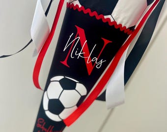 Peut inclure: Un cornet scolaire conique orné d'un motif de ballon de football noir et blanc. La section noire affiche le prénom Niklas en écriture rouge et blanche, un ballon de football, et le texte Schulanfang 11.08.2025, agrémenté de rubans rouges.