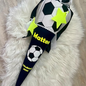 Könnte beinhalten: Ein blauer und schwarzer Schultüte mit einem Fußball-Design. Die Tüte trägt den Namen "Matteo" und den Text "Schulkind 2024". Es befinden sich zwei gelbe Sterne auf der Tüte.