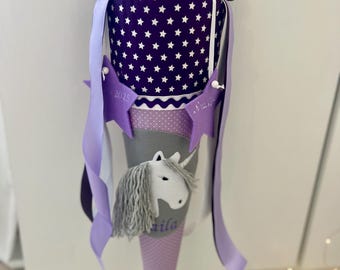 May include: Purple and gray school cone with a white unicorn design and the name "Mila" embroidered on the front. The cone is decorated with white stars and a purple ribbon.