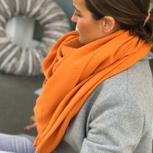 Op de afbeelding: Een vrouw draagt een grijze trui en een feloranje sjaal. De sjaal is gemaakt van een zachte, textuurrijke stof.