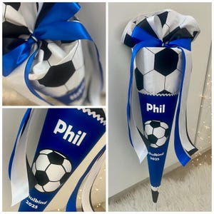 Könnte beinhalten: Eine blau-weiße Schultüte im Fußball-Design mit Fußballmuster und blauem Band. Auf der Tüte steht der Name "Phil" und der Text "Schulkind 2025".
