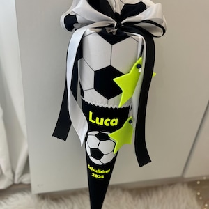 Könnte beinhalten: Eine schwarz-weiß Fußball-Geschenktüte in Kegelform mit einer großen Schleife. Die Tasche hat ein Fußball-Design und den Namen "Luca" in Gelb. Zwei limettengrüne sternförmige Dekorationen sind angebracht.
