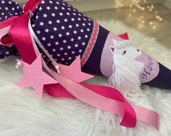 May include: A purple and white star-patterned school cone with a pink ribbon and star decorations. The cone features a unicorn design with the name "Hannah" written on it. The cone is designed for children.
