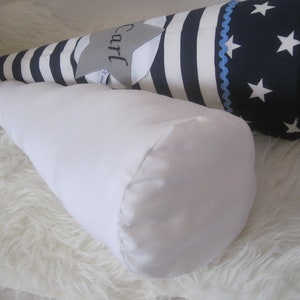 Könnte beinhalten: Ein weißes, zylindrisches Kissen mit einem blau-weiß gestreiften Stoffkegel mit einem Stern und dem Namen "Carl" darauf. Der Kegel und das Kissen liegen auf einer weißen, flauschigen Oberfläche.