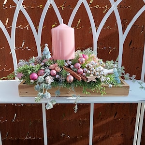 Peut inclure: Centre de table de Noël dans un plateau en bois, avec une grande bougie rose, des ornements et de la verdure. L'arrangement comprend des boules roses et argentées, une cannelle et de petits arbres décoratifs. Présenté sur une étagère en métal blanc.