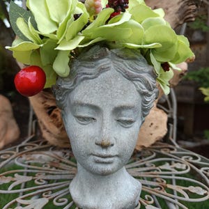 Op de afbeelding: Een grijze stenen hoofdsulptuur met gesloten ogen, versierd met lichtgroene kunstbloemen, rode bessen en groene bladeren. De sculptuur staat op een decoratieve metalen standaard, een uniek tuinornament.