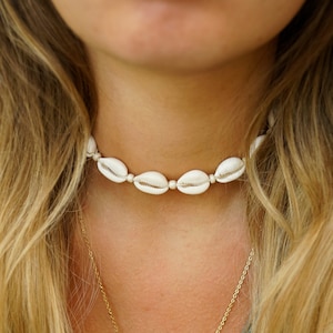 Könnte beinhalten: Eine Frau trägt eine weiße Muschel-Choker-Kette und eine goldene Kettenkette mit einem Muschel-Anhänger.