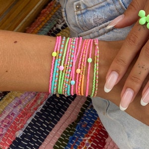Puede incluir: Un conjunto de pulseras de cuentas de colores, en rosa, verde, azul y amarillo, con pequeñas cuentas de colores. Las pulseras están apiladas en la muñeca de una persona.