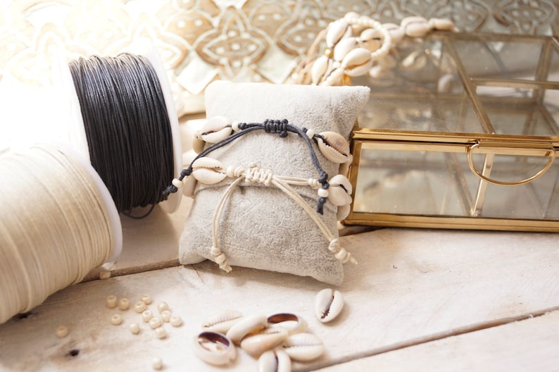 Puede incluir: Dos pulseras hechas con cord&oacute;n blanco y negro y conchas de cauri sobre un coj&iacute;n de terciopelo blanco. Hay carretes de cord&oacute;n blanco y negro y conchas de cauri sueltas sobre una superficie de madera blanca.