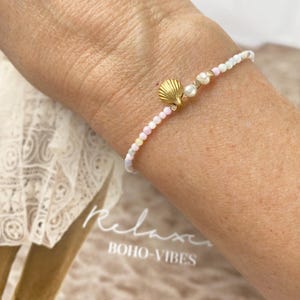Könnte beinhalten: Zartes Perlenarmband mit einem goldenen Muschel-Anhänger, kleinen Perlen und pastellfarbenen Perlen. Das Armband wird am Handgelenk getragen und zeigt das Design des Schmucks. Der Hintergrund enthält den Text "Relax Boho-Vibes".