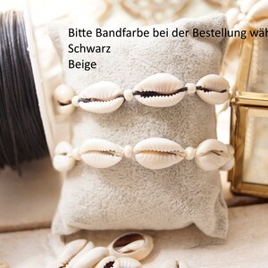 Puede incluir: Dos pulseras de conchas blancas con cord&oacute;n negro, expuestas sobre un coj&iacute;n de terciopelo beige. El texto "Bitte Bandfarbe bei der Bestellung w&auml;hlen: Schwarz Beige" es visible sobre las pulseras.
