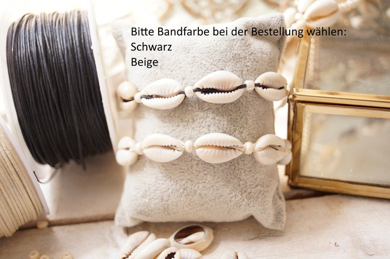 Puede incluir: Dos pulseras de conchas blancas con cord&oacute;n negro, expuestas sobre un coj&iacute;n de terciopelo beige. El texto "Bitte Bandfarbe bei der Bestellung w&auml;hlen: Schwarz Beige" es visible sobre las pulseras.