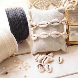 Puede incluir: Dos carretes de cord&oacute;n, uno negro y otro blanco, con conchas de cauri blancas y cuentas sobre una superficie de madera blanca.