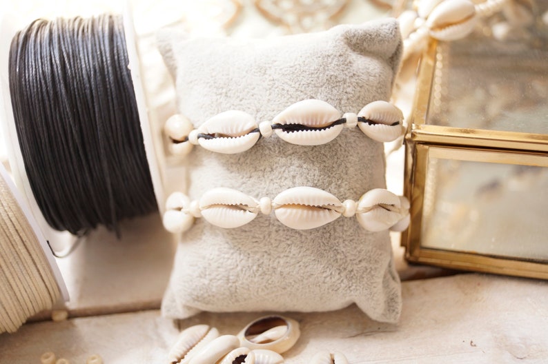 Puede incluir: Dos pulseras de conchas blancas con cord&oacute;n negro sobre un coj&iacute;n de terciopelo gris. Las pulseras est&aacute;n hechas con conchas de cauri y se muestran con carretes de cord&oacute;n negro y blanco.