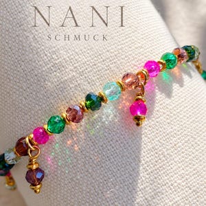Puede incluir: Una pulsera de cuentas con cristales facetados multicolores en tonos rosa, verde, azul y marrón, acentuada con espaciadores de metal dorado y dos dijes de cuentas colgantes. El texto HANDMADE BY NANI SCHMUCK es visible.