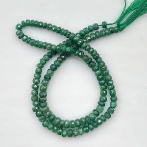 Peut inclure: Un collier de petites perles vertes facettées et rondes disposées en boucle circulaire. Les perles présentent des nuances variées de vert profond et de vert forêt avec une surface polie et réfléchissante. Un pompon en fil vert est attaché.
