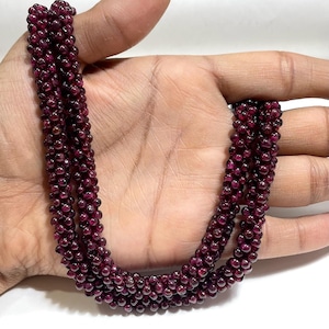 May include: A multi-strand necklace composed of numerous small, deep red, spherical garnet beads. The beads are arranged in a dense, textured pattern, creating a continuous, flexible rope-like structure that displays a consistent dark crimson hue.