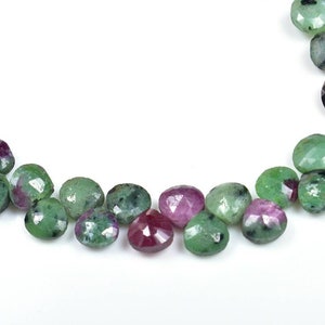 Pode incluir: Um fio de contas de pedras preciosas facetadas verdes e rosadas. As contas são em forma de coração e têm um acabamento polido.