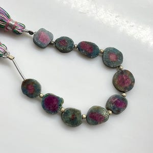 May include: A necklace featuring irregularly shaped, polished gemstones with a mix of teal and magenta hues. The stones are strung together with small silver beads. A colorful tassel with pink, green, and purple threads accents the necklace.