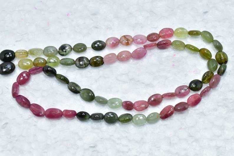 May include: A strand of oval-shaped gemstone beads in shades of pink, green, and black. The beads are strung on a clear thread and are arranged in a graduated pattern.