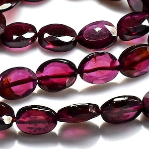 Könnte beinhalten: Eine Strähne ovaler, facettierter Granatperlen in einer tiefen, satten roten Farbe. Die Perlen sind auf einem durchsichtigen Faden aufgefädelt und in einem lockeren, fließenden Muster angeordnet.