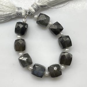 Op de afbeelding: Een streng van gefacetteerde, donkergrijze, kubusvormige kralen afgewisseld met kleine, ronde, witte parels. De streng is aan de bovenkant samengebonden met een zilverkleurige metalen ring en twee lange, witte zijden kwastjes met zilverdraad.