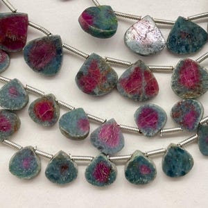 May include: Strands of gemstone beads with a teardrop shape. The beads have a mix of colors, including deep red and teal. Each bead is strung on a silver-colored wire, creating a necklace or bracelet.