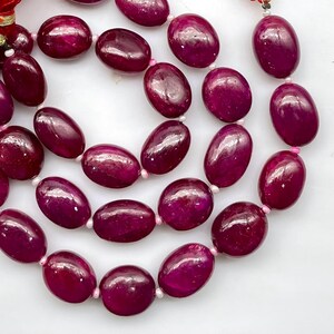 May include: A strand of smooth, oval-shaped deep magenta ruby beads arranged in a curved pattern. Each polished gemstone is separated by small, light pink spacer beads, creating a continuous string of vibrant, dark pink jewelry components.