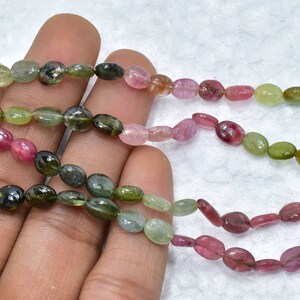 May include: A strand of oval-shaped, multi-colored gemstone beads. The beads are shades of green, pink, and black.