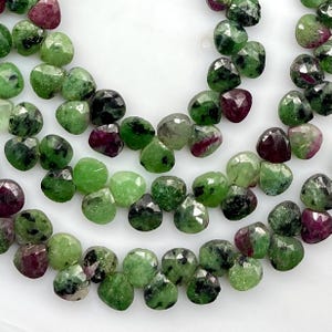 Puede incluir: Primer plano de una colección de cuentas de piedras preciosas facetadas. Las cuentas son una mezcla de colores verde y rojo intenso. Las piedras preciosas tienen forma de corazón y parecen estar ensartadas. El fondo es blanco.