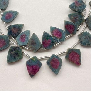 May include: Strands of gemstone beads. Each bead is a shield shape with a teal-green exterior and a vibrant magenta center. The beads are strung on a silver-colored wire, creating a necklace or bracelet component. The gemstones have a natural, organic appearance.