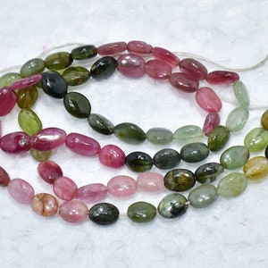May include: A strand of oval-shaped gemstone beads in shades of pink, green, and black. The beads are strung on a white thread.