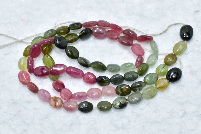 May include: A strand of oval-shaped gemstone beads in shades of pink, green, and black. The beads are strung on a white thread.