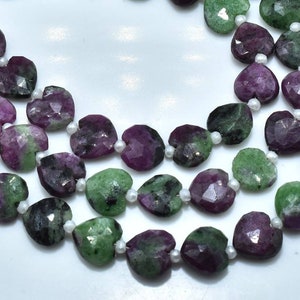 May include: Close-up of heart-shaped gemstone beads in green and purple hues, with small white beads between them. The faceted cut of the beads creates a shimmering effect. The beads are strung together, possibly for a necklace or bracelet.