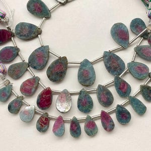 May include: Strands of gemstone beads featuring teardrop-shaped stones. The stones have a teal-blue hue with a central splash of magenta. The beads are strung on a silver-colored wire, creating a delicate and elegant look. These beads are ideal for jewelry making.