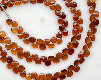May include: A strand of faceted, heart-shaped garnet beads in deep shades of orange and red. The translucent gemstones are strung together on a thin wire, showcasing a polished, reflective surface and a consistent, uniform cut throughout the strand.