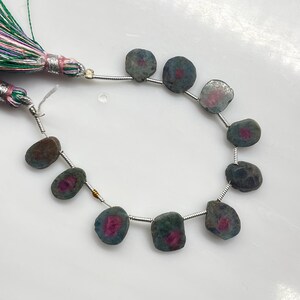 May include: Strand of natural gemstone beads. The beads are a mix of dark green, gray, and brown with a vibrant pink center. The beads are strung on a silver wire, and a tassel with green, pink, and silver threads is attached.