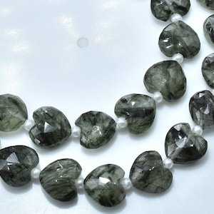 May include: Two strands of heart-shaped beads. The beads are a translucent, smoky green color with dark inclusions, giving them a unique, textured appearance. Each heart is faceted, and strung with small white beads.