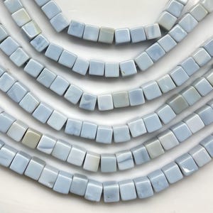 Puede incluir: Primer plano de varios hilos de cuentas cuadradas de color azul claro. Las cuentas tienen un aspecto ligeramente veteado, con variaciones de color que van del azul pálido al blanquecino. Las cuentas están ensartadas, creando un patrón curvo y fluido.